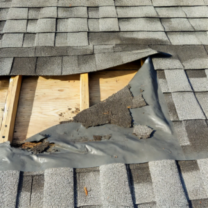 A close-up image of a damaged roof. The shingles are missing in a large area, exposing the roof deck and underlayment.